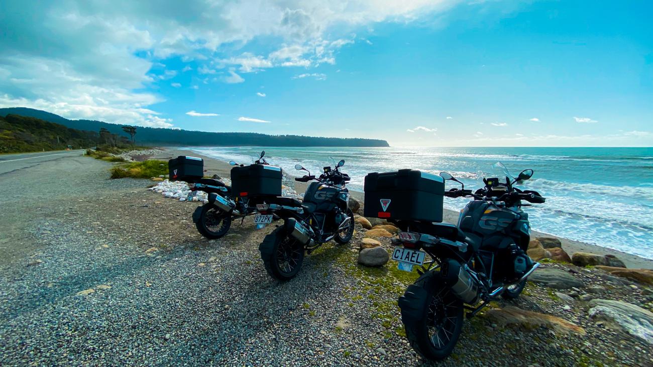 Three BMW GS motorcycles parked on a rocky beach along New Zealand's rugged west coast with waves breaking in the background.