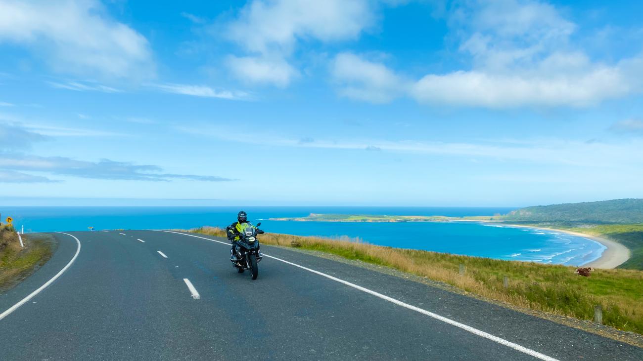 Motorcyclist riding along a sweeping New Zealand coastal highway with turquoise ocean bay and green hills in the background.