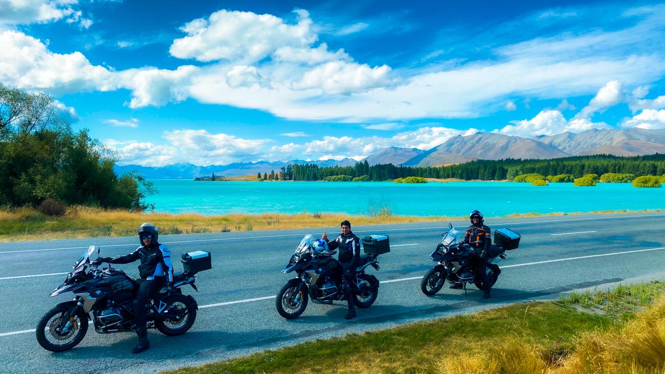 Three motorcyclists parked beside the stunning turquoise waters of Lake Tekapo with snow-capped mountains in the distance.