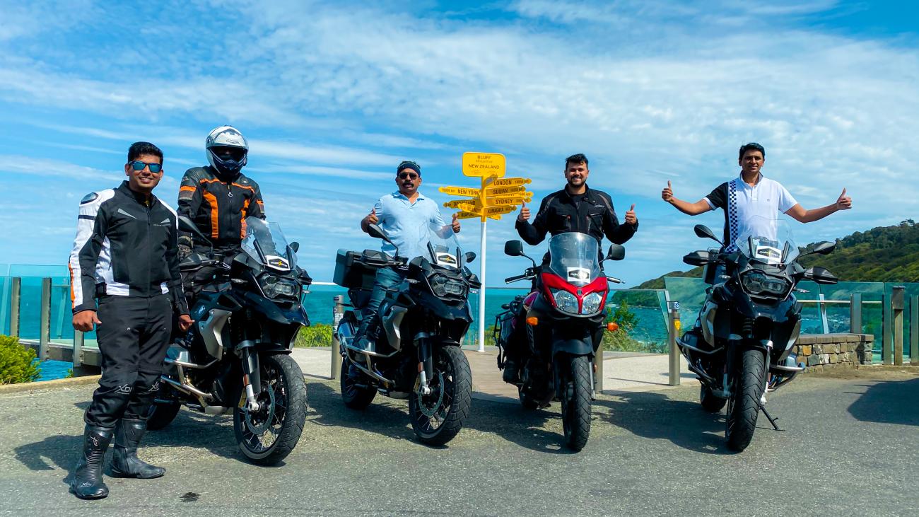 Four riders with their motorcycles posing at the famous Bluff signpost in New Zealand showing distances to world cities.