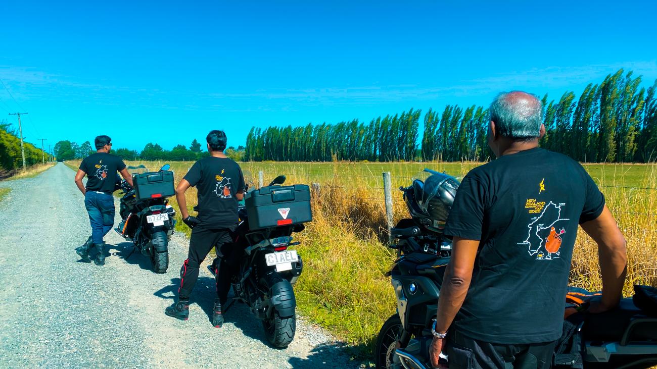 Three riders in matching tour t-shirts standing with their motorcycles on a gravel road lined with poplar trees in rural New Zealand.