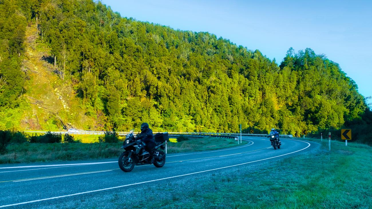 Two motorcyclists riding through a winding road surrounded by lush green native forest on New Zealand's South Island.