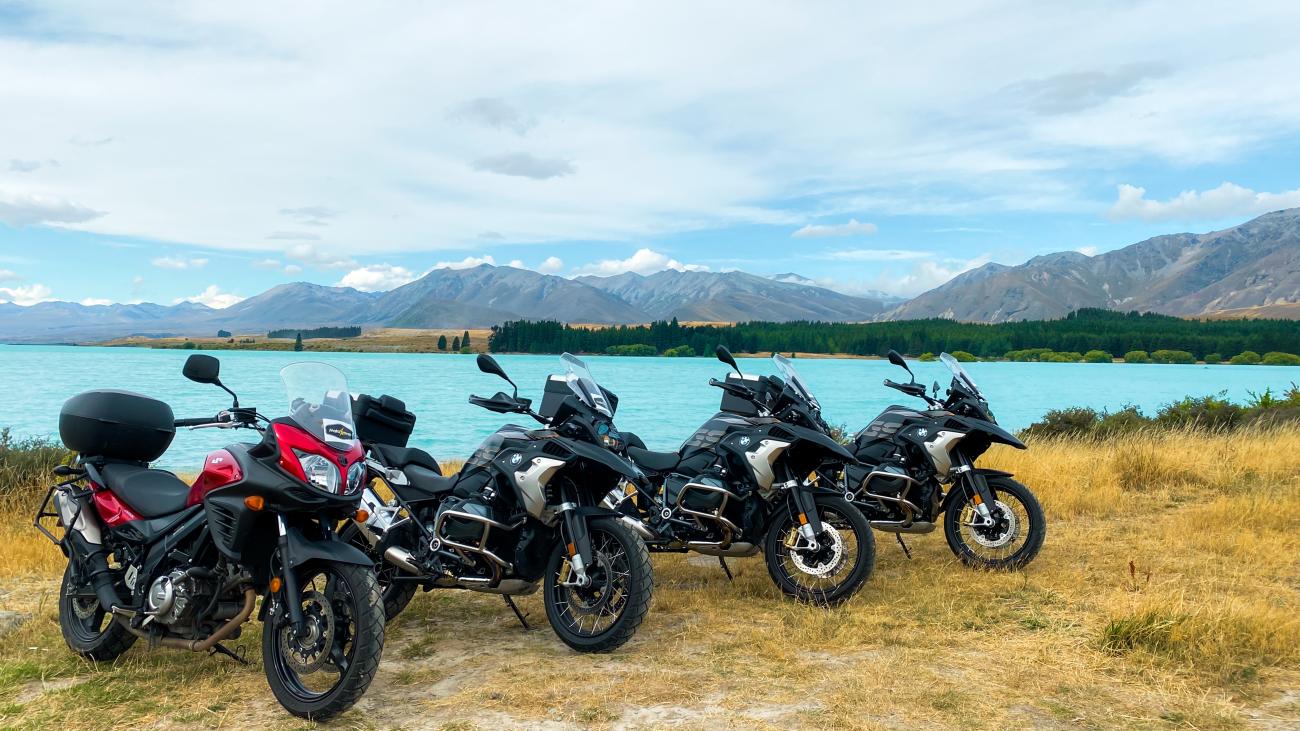 Four touring motorcycles lined up by the turquoise Lake Tekapo with the Southern Alps mountain range behind.