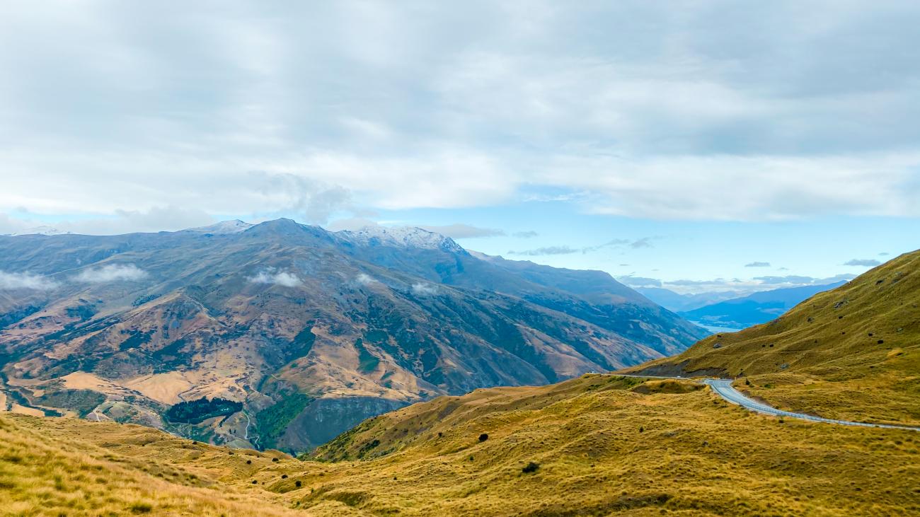 Winding Crown Range Road climbing through golden alpine grasslands with snow-dusted mountains in the distance.