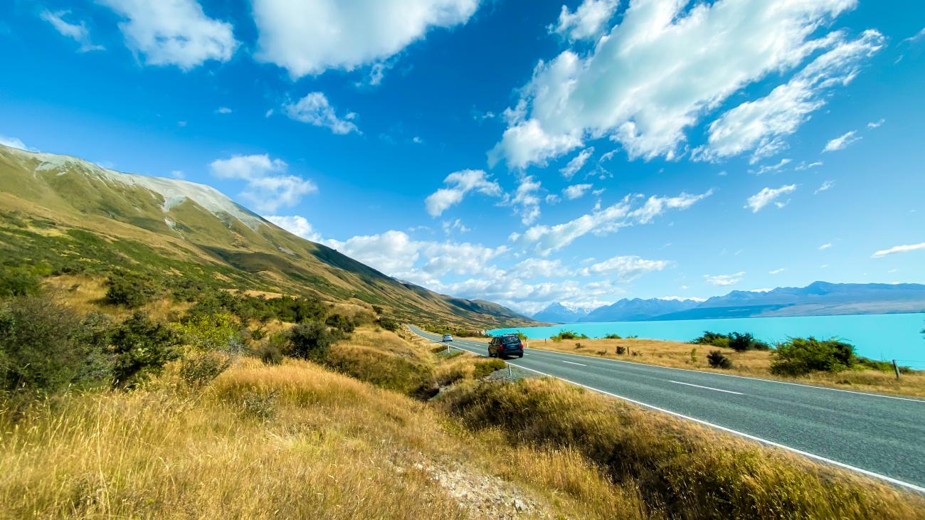 Car driving along a scenic highway beside turquoise Lake Pukaki with snow-capped mountains in the distance.