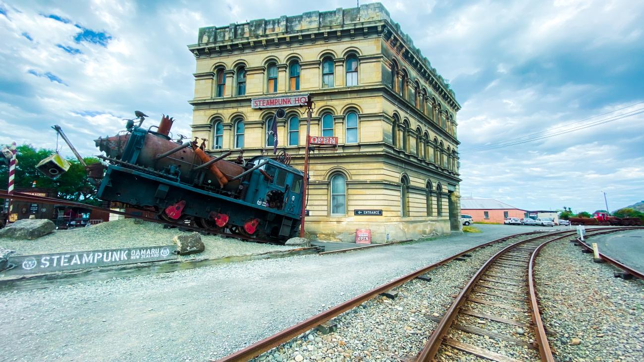 Steampunk HQ museum in Oamaru with a vintage locomotive sculpture and railway tracks in front of the historic building.