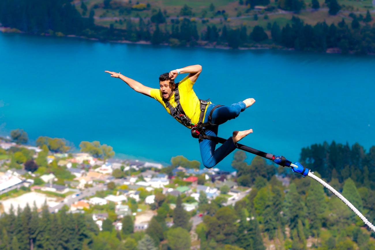 Man bungee jumping high above Queenstown with turquoise Lake Wakatipu and the town visible below.