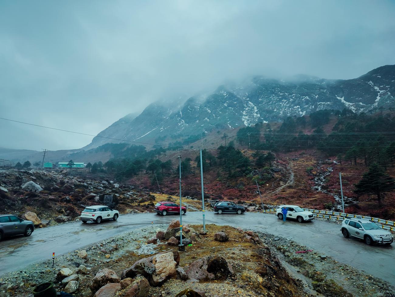 Cars parked at a high-altitude mountain pass with snow-dusted peaks and autumn-colored shrubs.