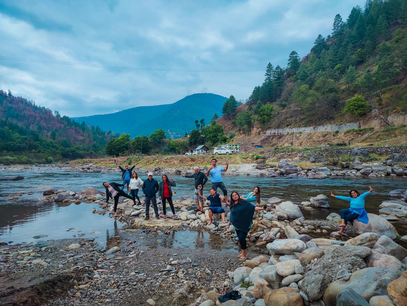 A group of travelers posing on rocks along a river with pine-covered mountains and an overcast sky.