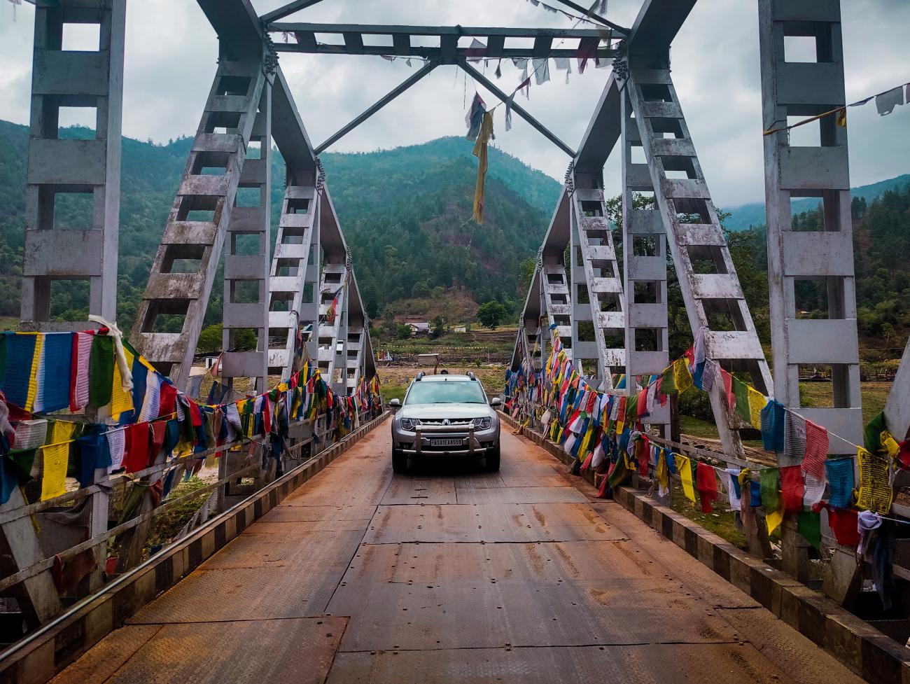 An SUV crossing a steel bridge decorated with colorful Buddhist prayer flags with green mountains behind.