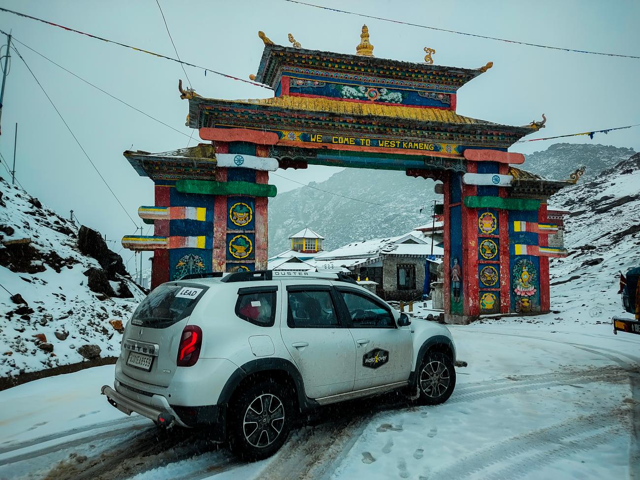 A white SUV passing through the ornate Sela Pass gateway in West Kameng with snow covering the ground.