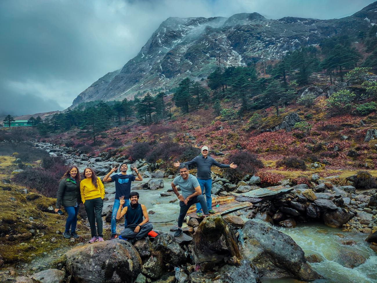 Travelers posing by an alpine stream with snow-capped peaks and colorful highland vegetation near Tawang.
