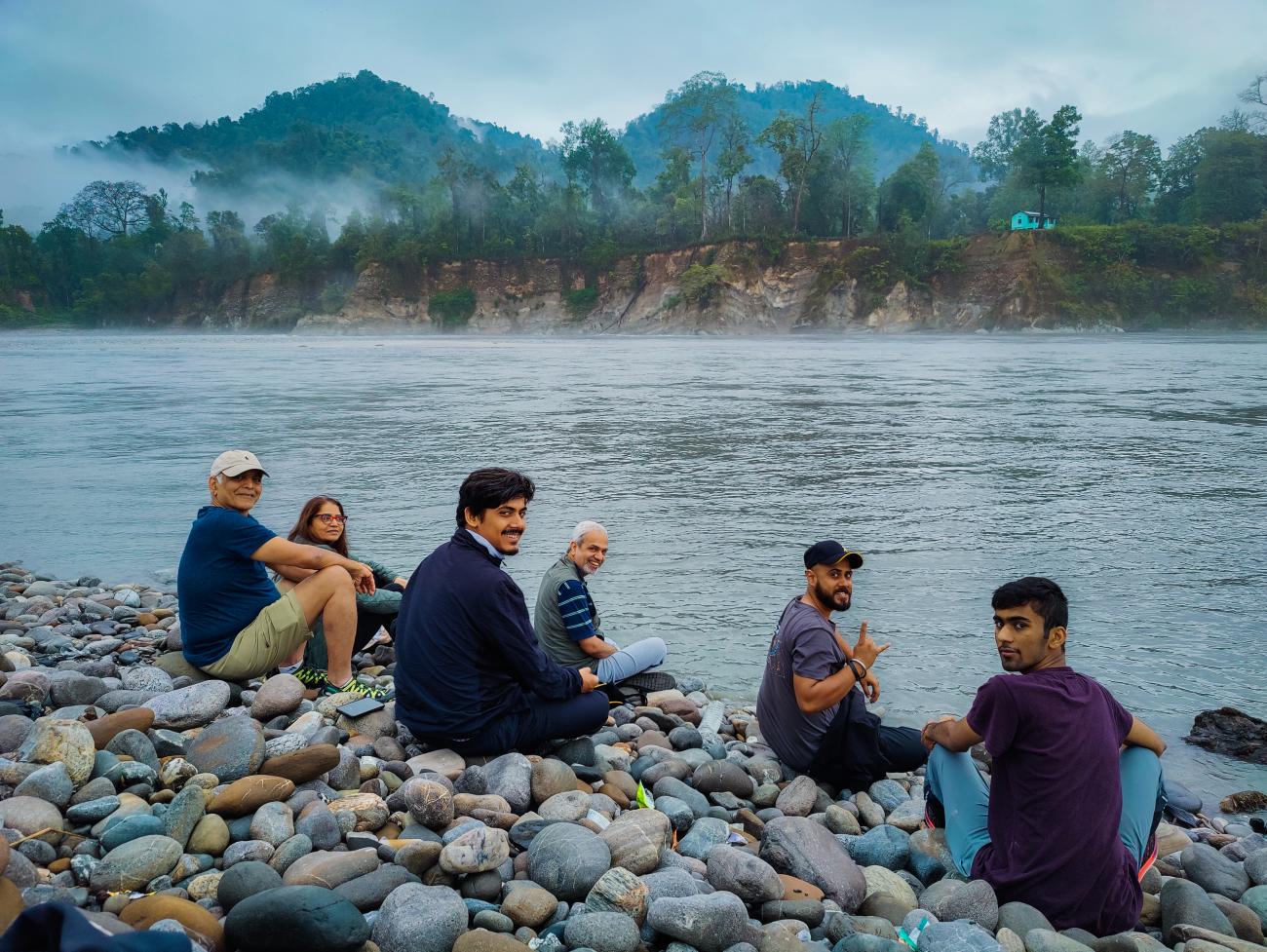Travelers sitting on a rocky riverbank with mist rising over the water and forested mountains in the background.
