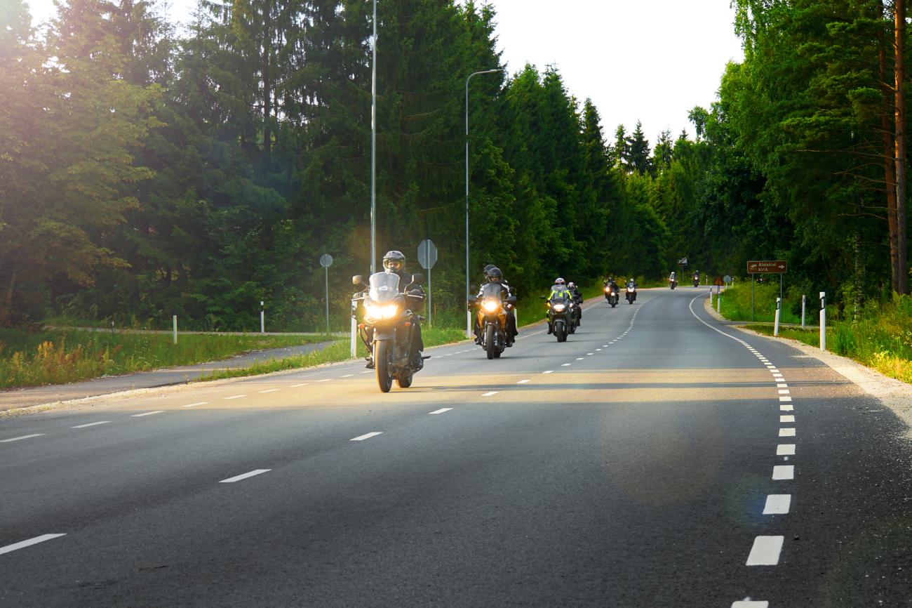 Convoy of motorcyclists riding through a tree-lined forest road in the Baltic region at golden hour.