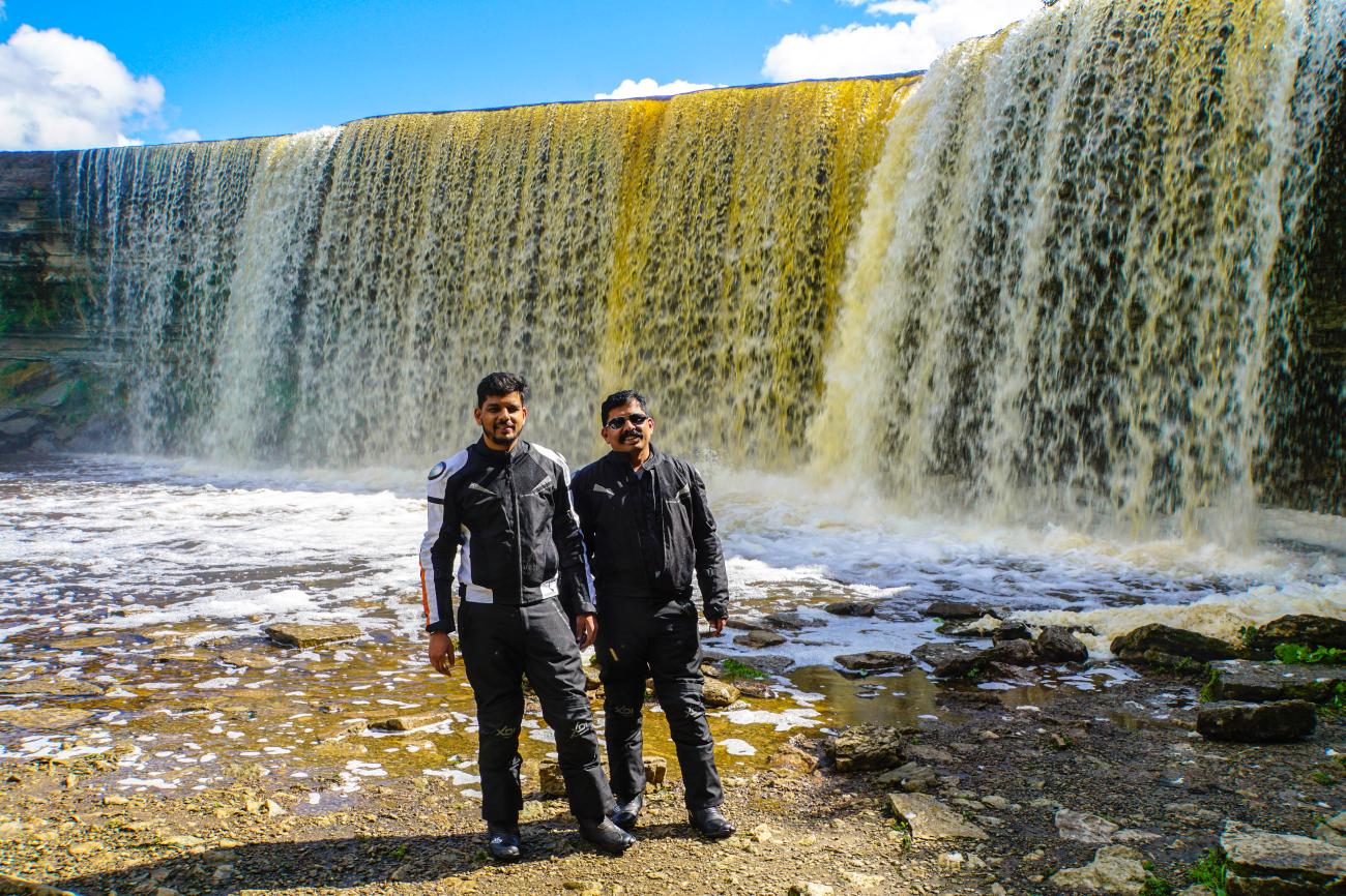 Two riders in motorcycle gear standing in front of a wide waterfall in Estonia.