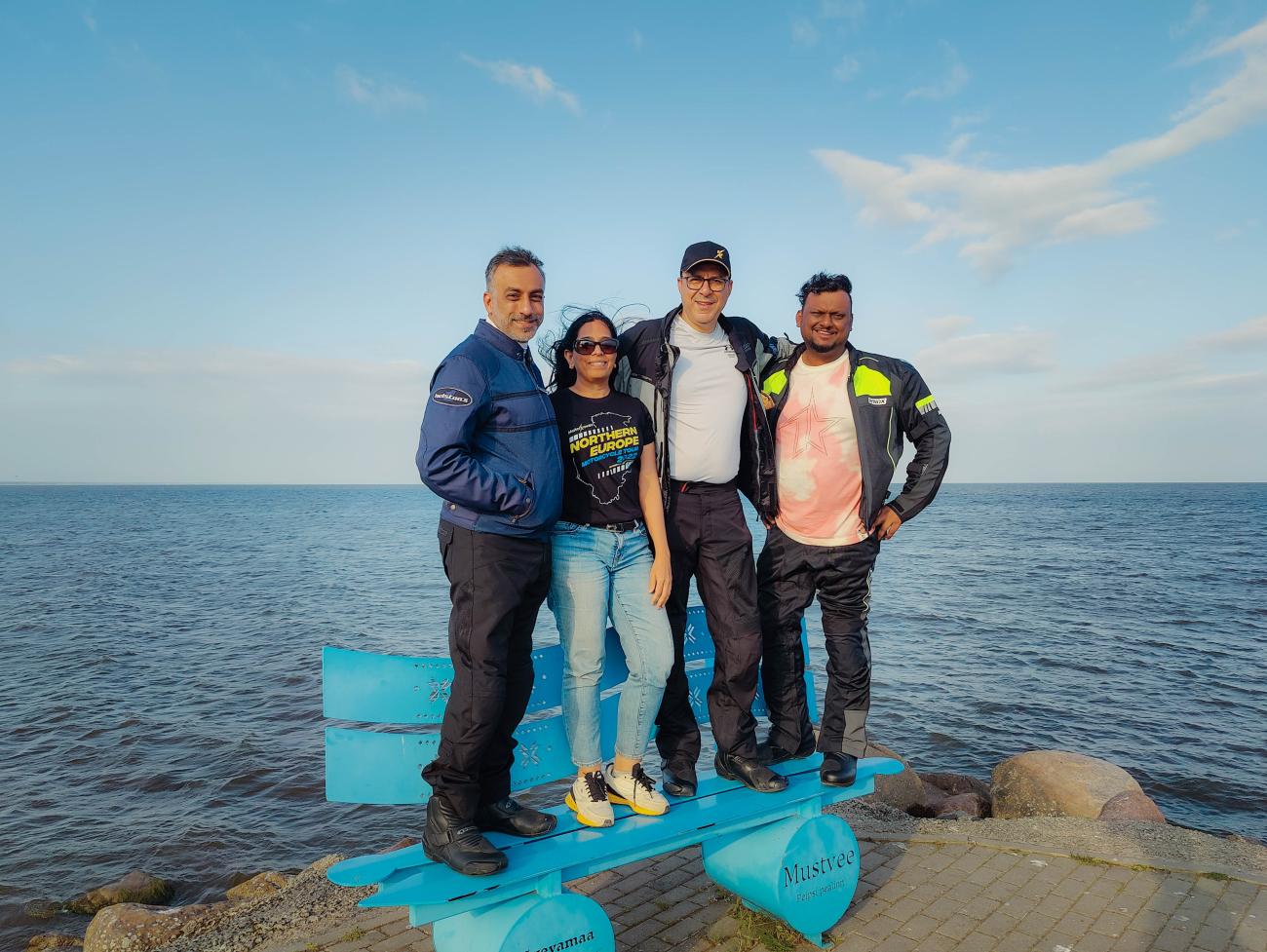 Four tour riders standing on a blue bench sculpture by the Baltic Sea near Mustvee, Estonia.
