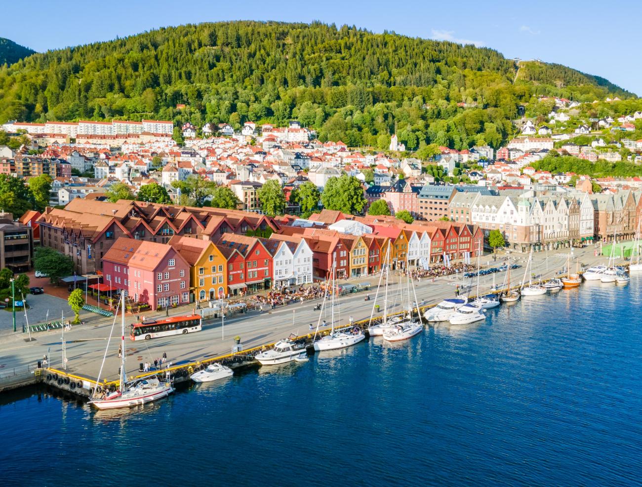 Aerial view of Bergen's colourful Bryggen waterfront with boats moored in the harbour and green hillside behind.