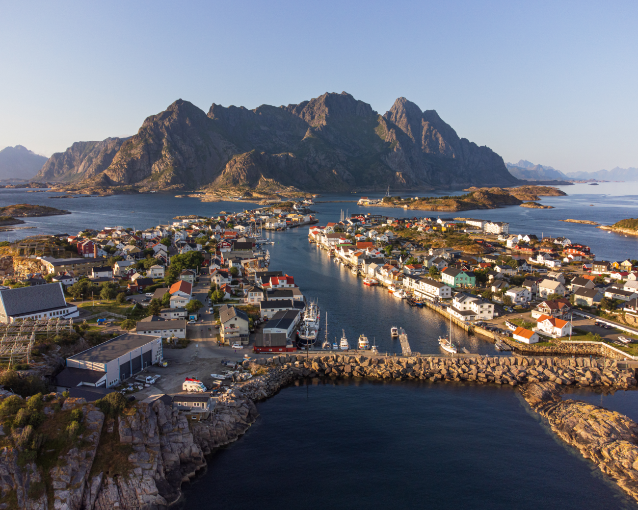 Aerial view of a Lofoten fishing village surrounded by dramatic peaks and calm harbour waters at golden hour.