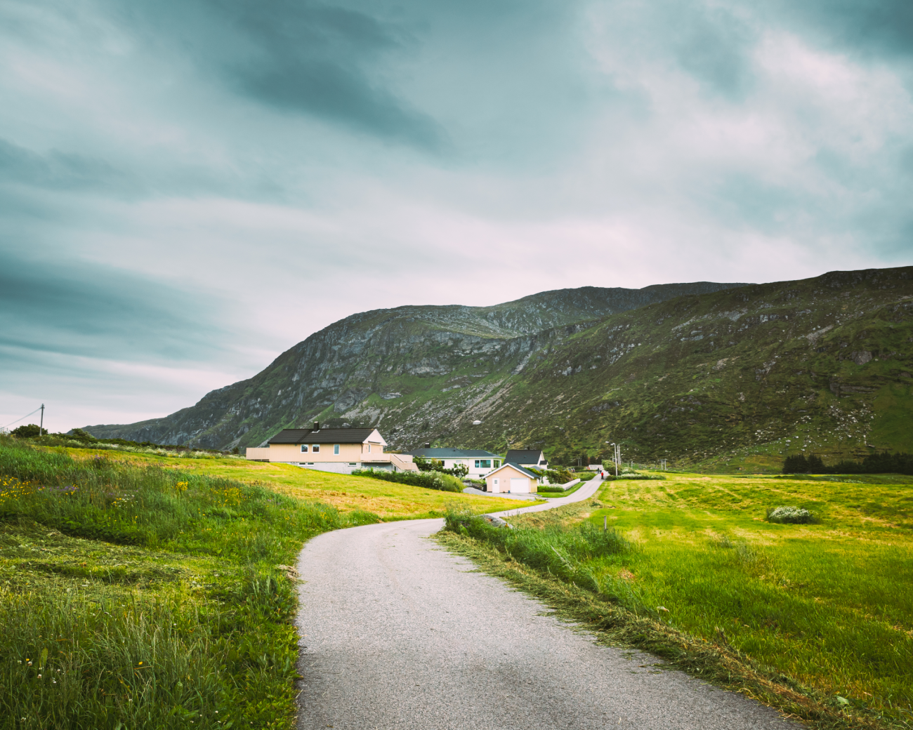 Narrow country road leading through a small Norwegian village surrounded by green hills under moody skies.