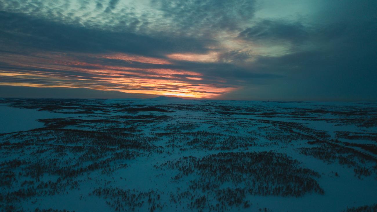 An aerial view of endless snow-covered taiga forest stretching to the horizon under a dramatic orange and teal sunset.