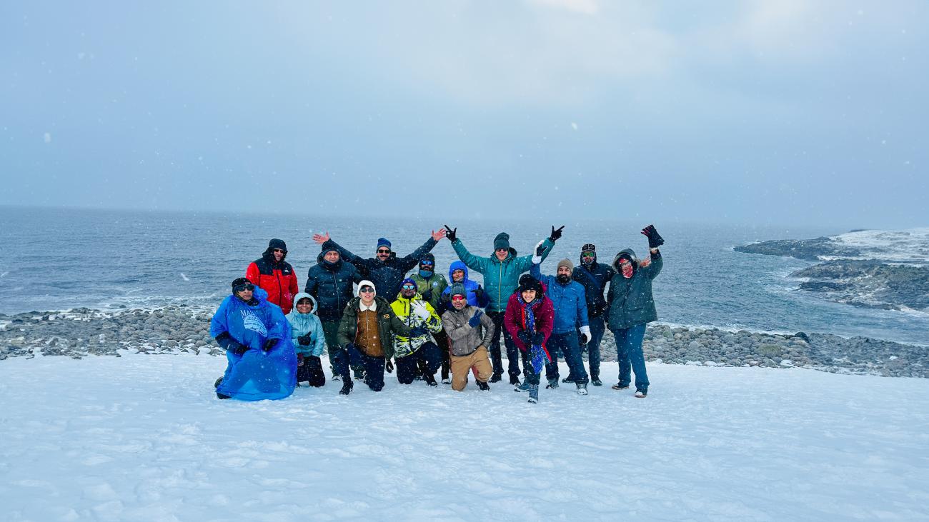 Tour group posing together on a snowy Arctic Ocean shoreline with waves crashing and snowflakes in the air.