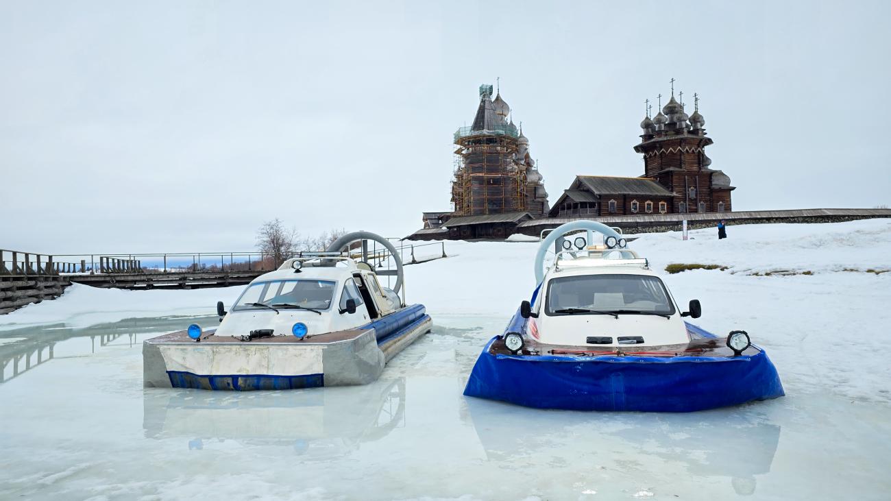 Two hovercrafts resting on a frozen lake with the wooden churches of Kizhi Island visible in the background.