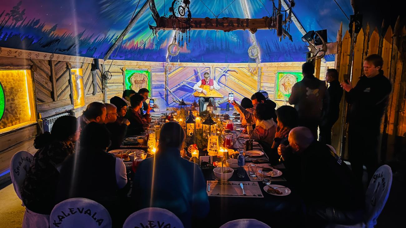 Tour guests seated at a candlelit dinner table inside a traditional Kalevala-themed wooden lodge with colorful northern lights murals.