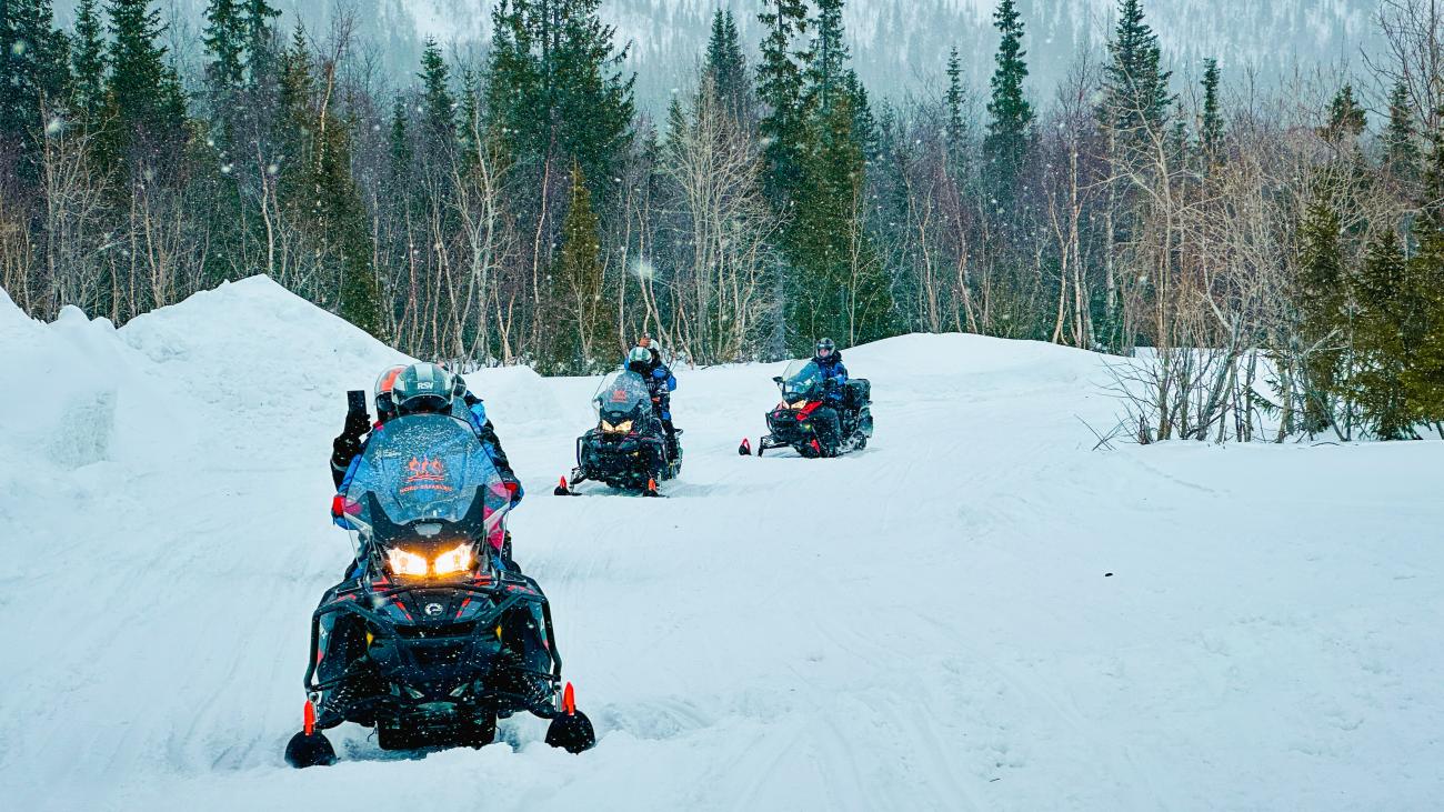 Three snowmobiles riding through deep snow in a Russian forest with headlights glowing and snowflakes falling.