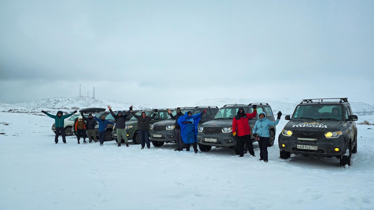 Tour participants cheering with arms raised alongside a row of MotoRover SUVs on a snow-covered tundra.