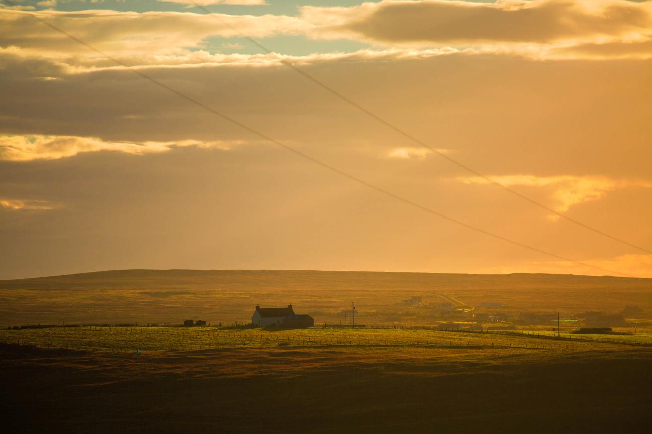 A lone stone farmhouse silhouetted against a golden sunset over the wide open moorland of northern Scotland.