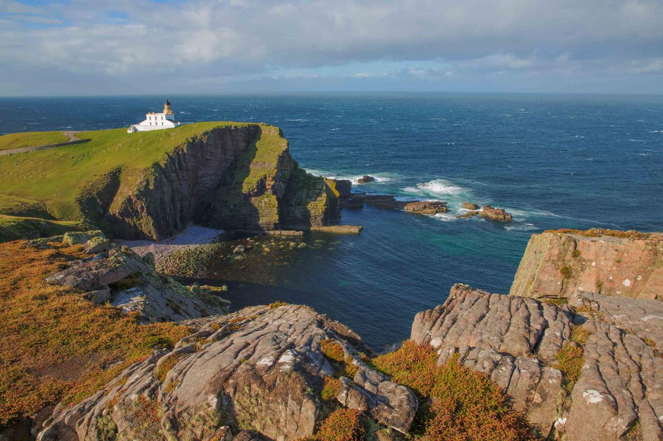 A white lighthouse perched on a grassy cliff edge above rugged sea stacks and crashing Atlantic waves on the Scottish coast.