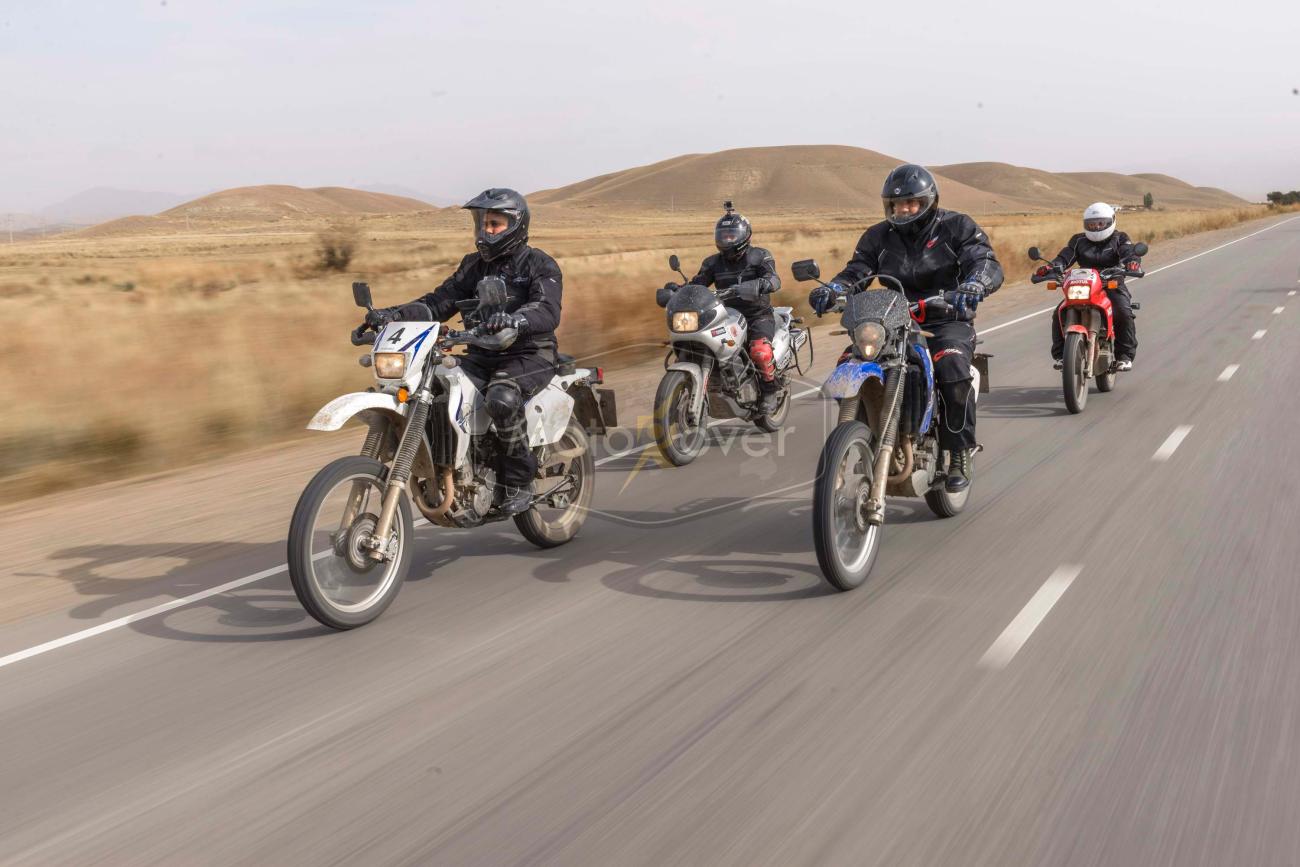 Four motorcyclists ride in formation on an open highway through rolling arid hills with a MotoRover watermark visible.