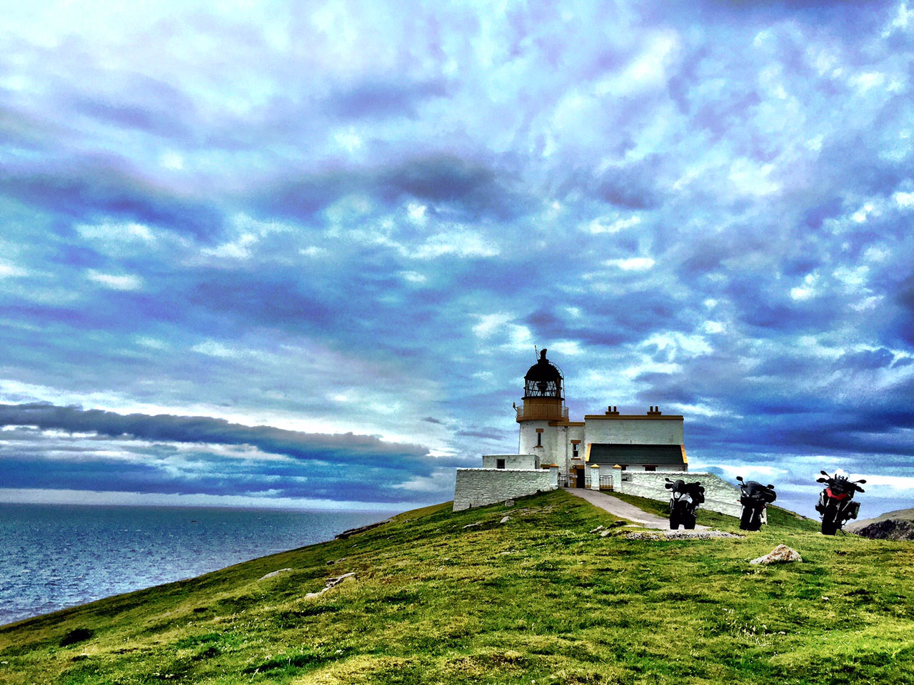 Two adventure motorcycles parked beside the white Stoer Head lighthouse on a grassy headland overlooking the Atlantic under a vivid cloudy sky.