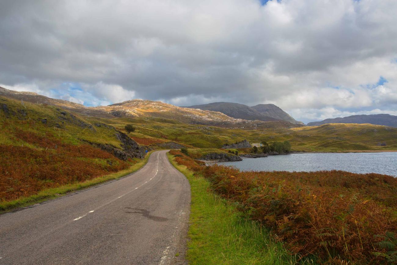 A winding single-track road curves alongside a Highland loch surrounded by golden bracken and heather-covered hills under a moody sky.