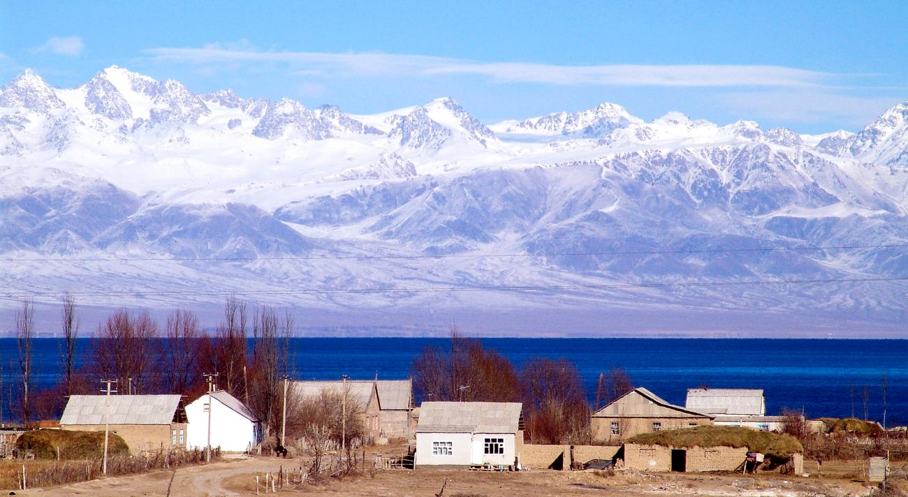 A lakeside village on Issyk-Kul with deep blue water and the snow-covered Tien Shan mountain range beyond.