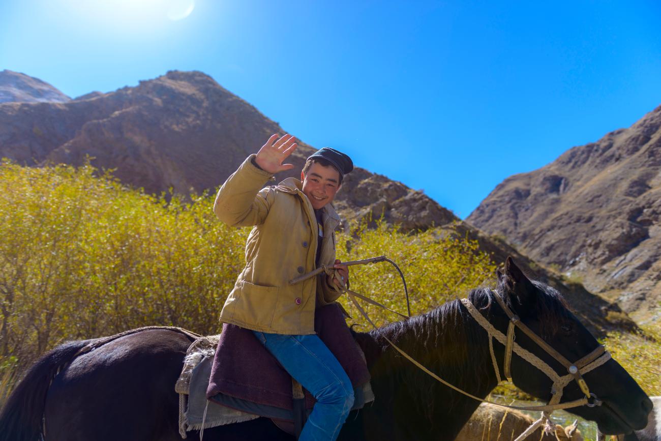 A young Kyrgyz boy on horseback waves and smiles in a rocky mountain valley with autumn foliage.