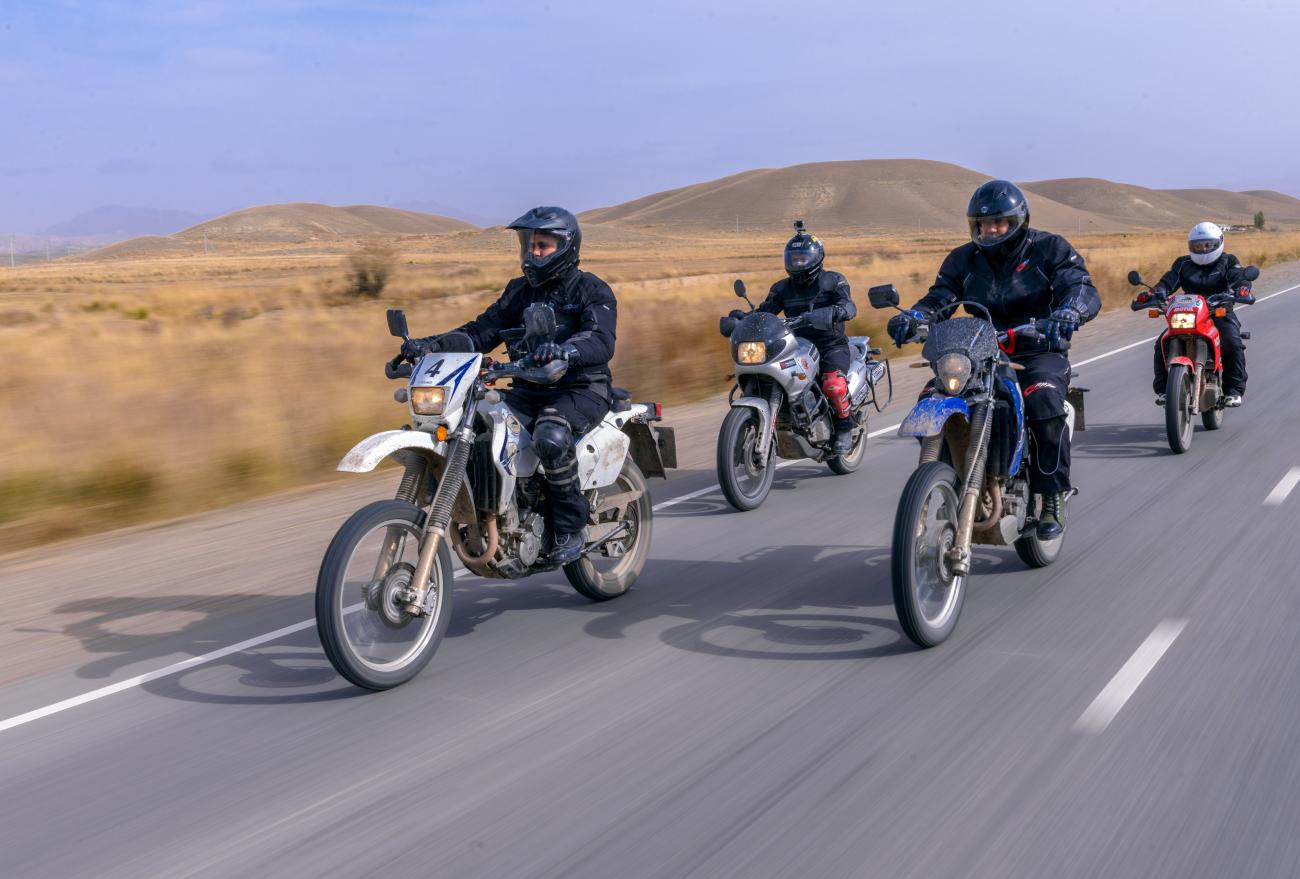 Four motorcyclists ride side by side along a steppe highway with rolling brown hills in the background.