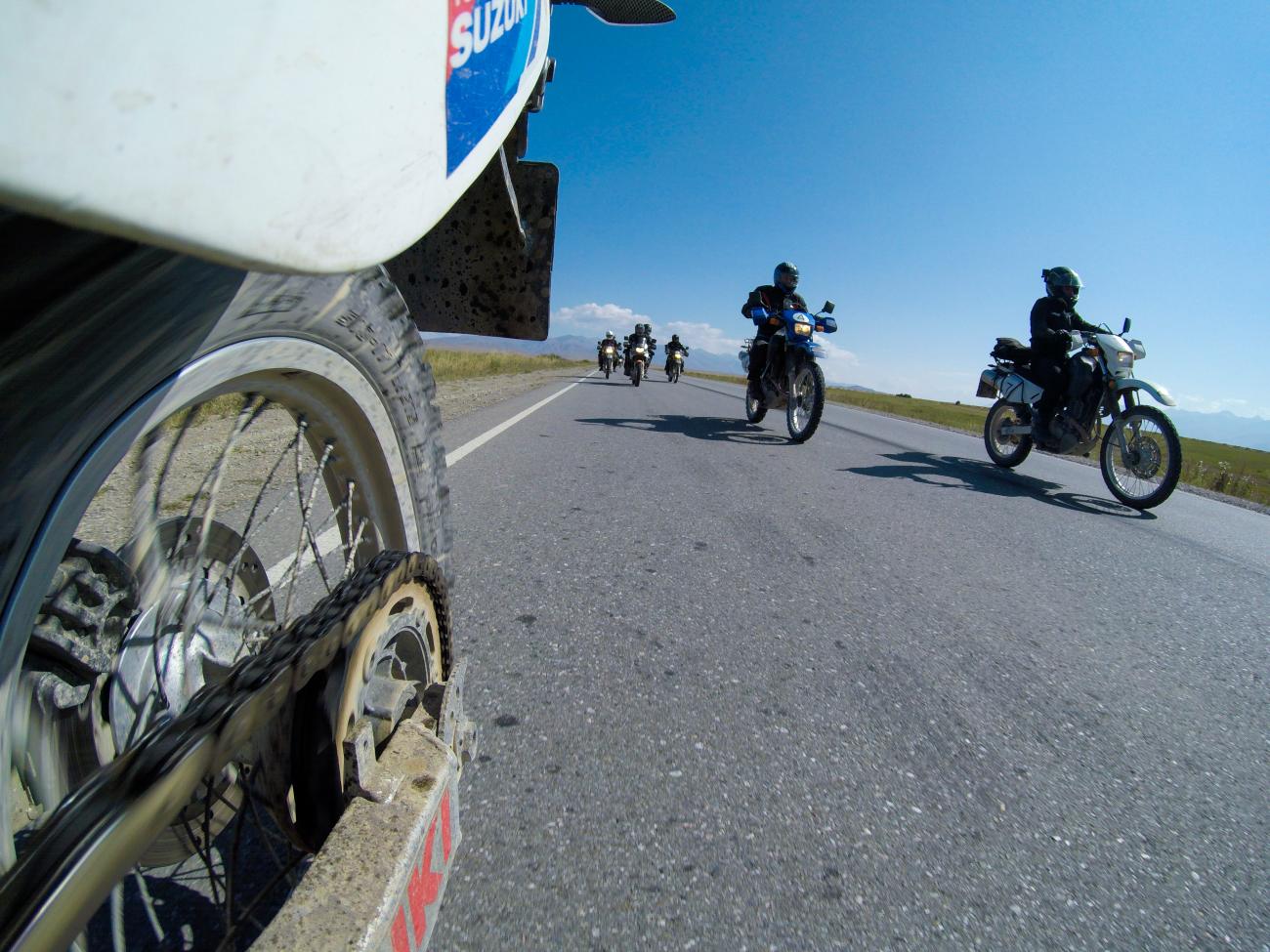 Low-angle shot from a motorcycle rear wheel showing fellow riders approaching on an open Central Asian road.