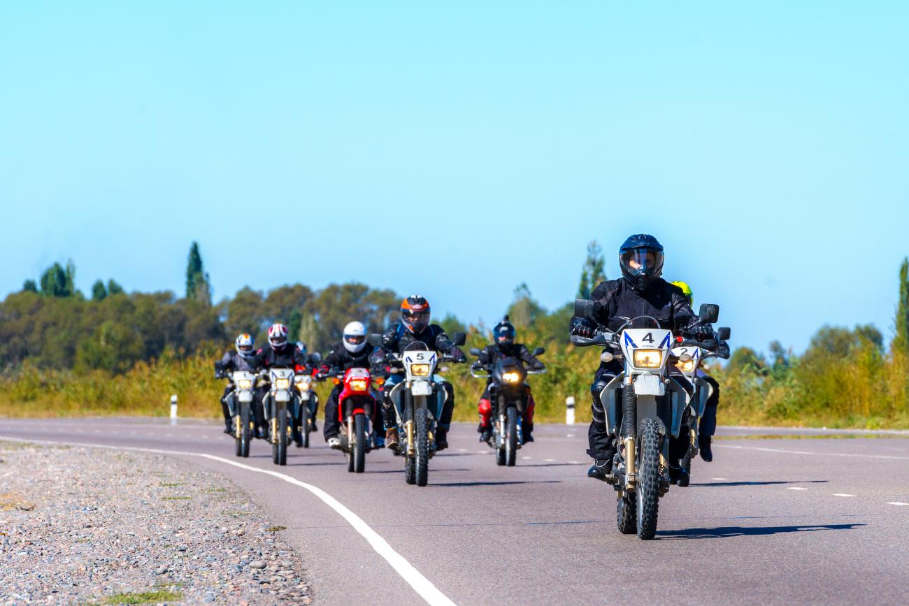 A group of adventure motorcyclists rides toward the camera on a paved road lined with greenery in Central Asia.
