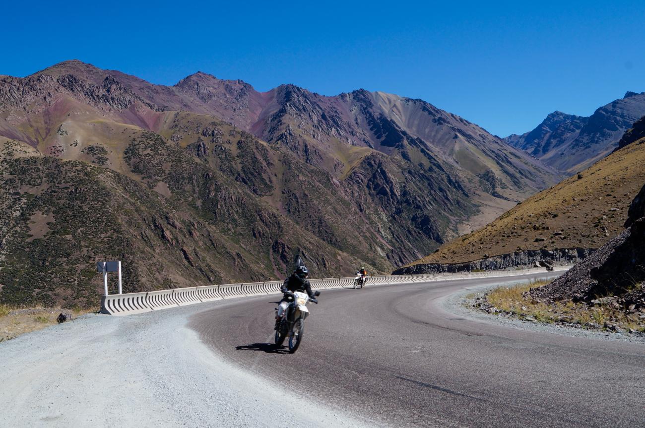 A motorcyclist navigates a hairpin bend on a high mountain pass with rugged multicolored peaks rising above.