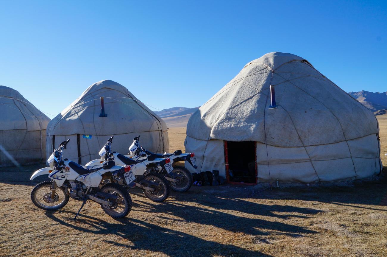 Adventure motorcycles parked beside traditional white yurts on an open plain with mountains in the distance.