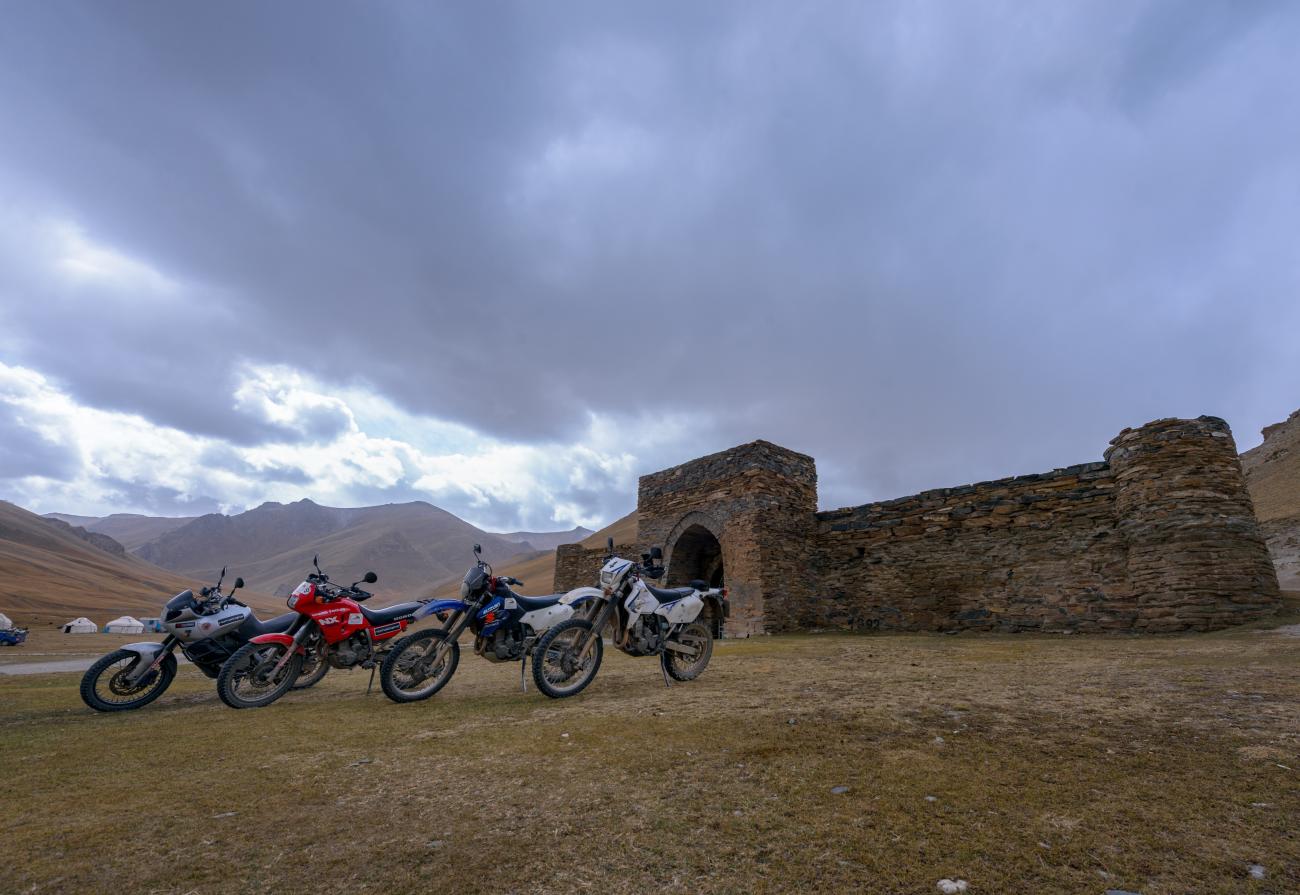 Three adventure motorcycles parked in front of the ancient stone Tash Rabat caravanserai with yurts and mountains in the background.