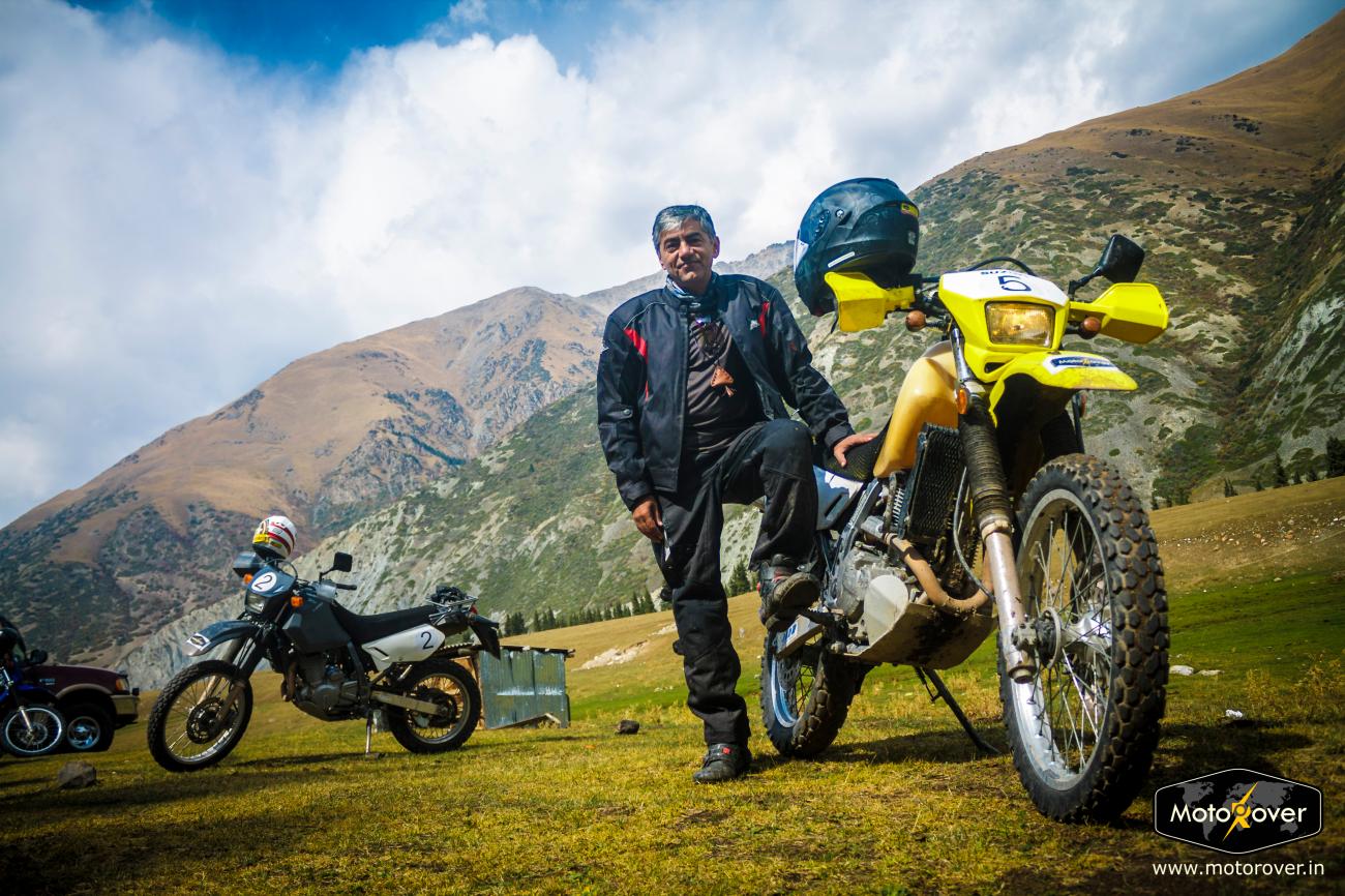 A rider stands beside his yellow adventure motorcycle on an alpine meadow with dramatic mountain peaks and cloudy skies.