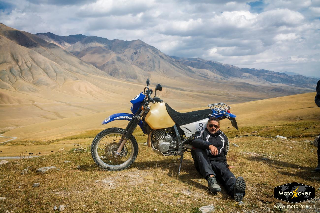 A rider relaxes on the ground beside his motorcycle at a high-altitude pass with sweeping barren mountain views.