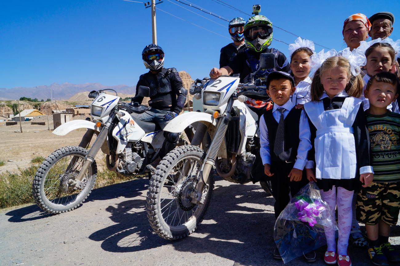 Two motorcyclists pose with a group of smiling local schoolchildren in a Kyrgyz village with arid hills behind.