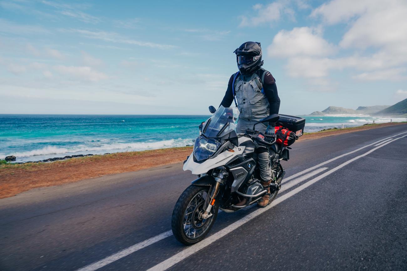 Motorcyclist on a BMW R 1250 GS riding along a South African coastal road with crashing ocean waves and mountains in the distance.