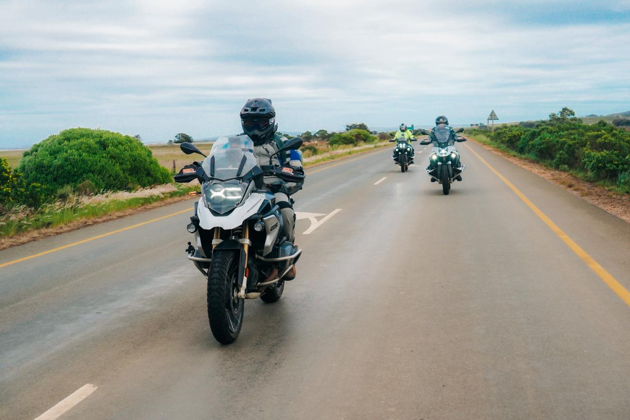 Group of BMW GS motorcyclists riding in formation on an open South African highway through green fynbos landscape.