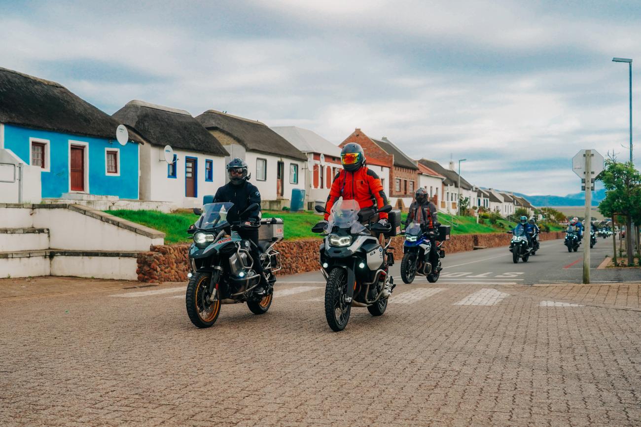 Motorcyclists riding BMW adventure bikes through a colourful South African village with traditional thatched-roof houses.