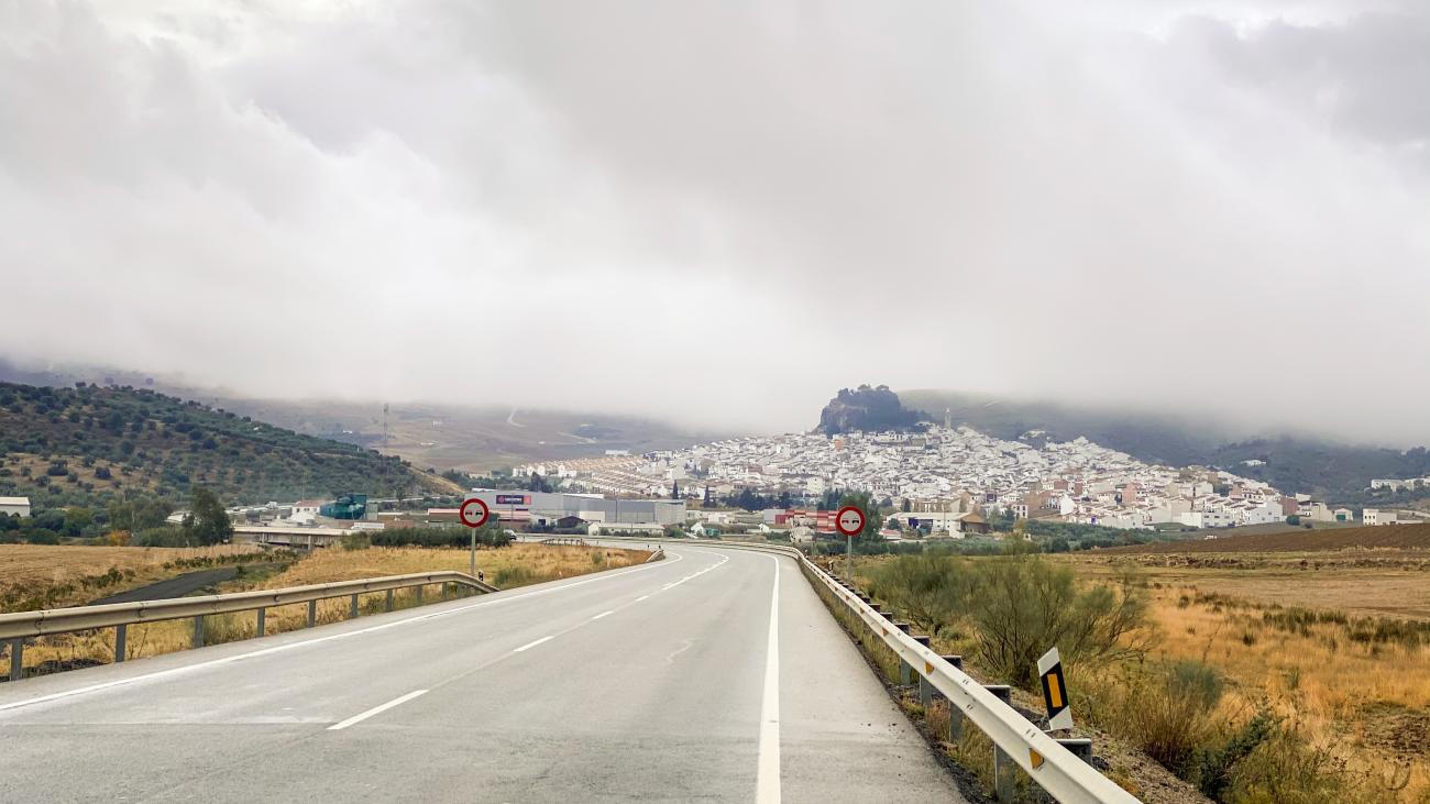 Open road leading toward a white hilltop village shrouded in low clouds in the Andalucian countryside.