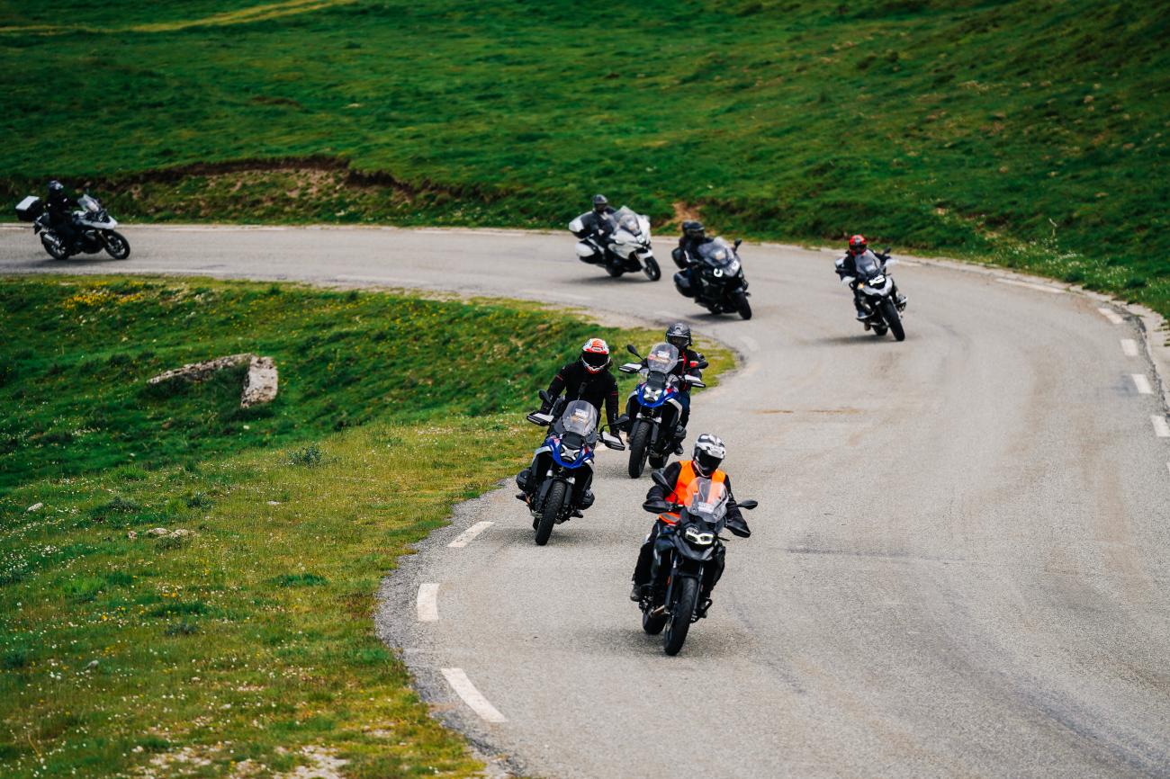 Group of motorcyclists climbing hairpin bends on a green Pyrenees mountain road viewed from above.